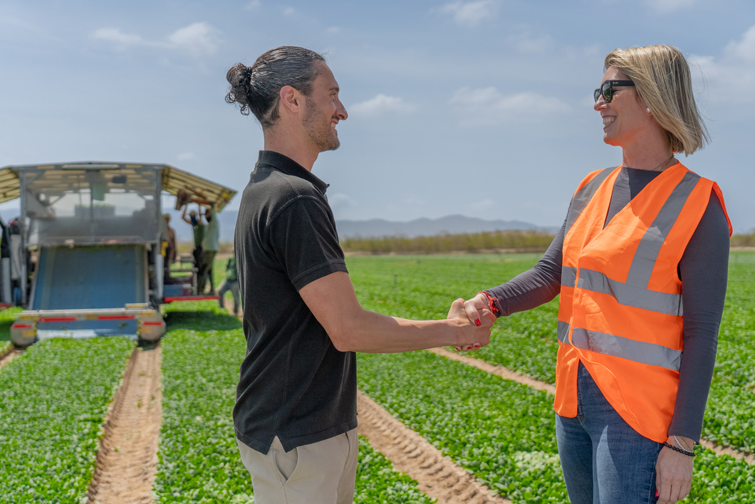A man and a woman shaking hands in a field, one wearing a high-visibility vest, with workers and harvesting equipment in the background, symbolising partnership and collaboration in agriculture.