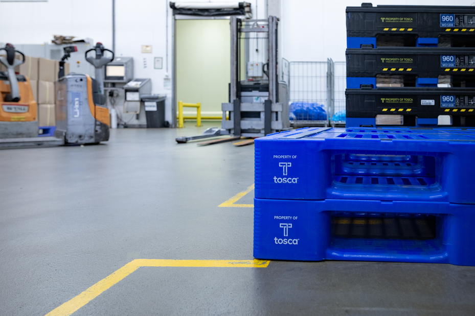 Two blue Tosca-branded reusable plastic pallets stacked in a warehouse aisle, with pallet jacks and industrial equipment visible in the background, highlighting durable pharmaceutical packaging solutions.