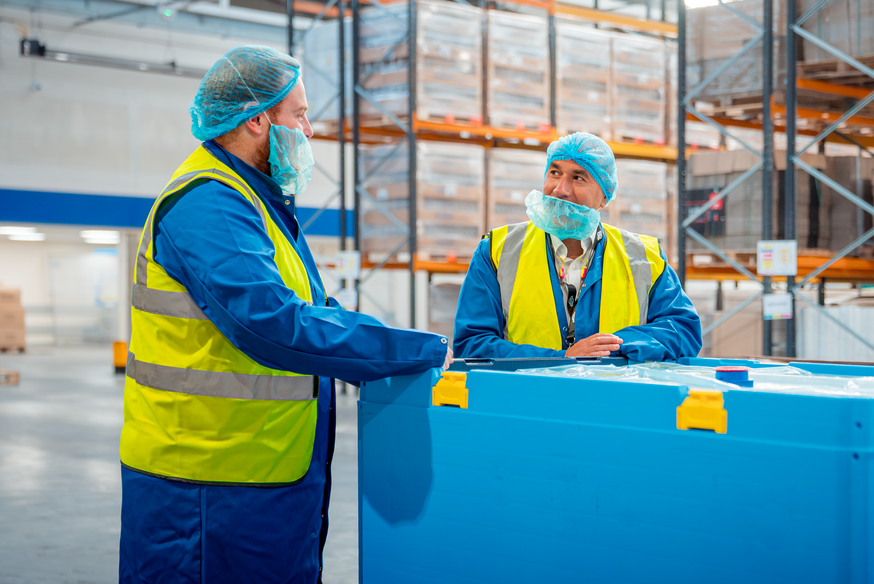 A customer and a client talk over a reusable plastic bulk bin, in a factory environment.