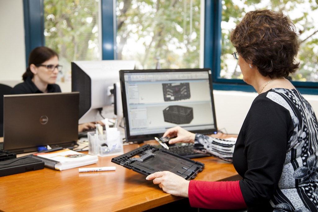 A research and development specialist holds a side panel of a plastic crate to check product specifications while comparing quality requirements displayed on a computer screen.