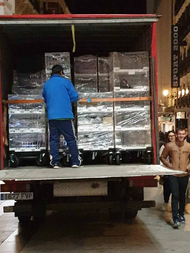 A warehouse worker unloads the back of a freight truck carrying Tosca Crystal Clear Crates filled with products stacked on portable dollies. These reusable plastic crates help maximise textile stock management and promote labour efficiency.