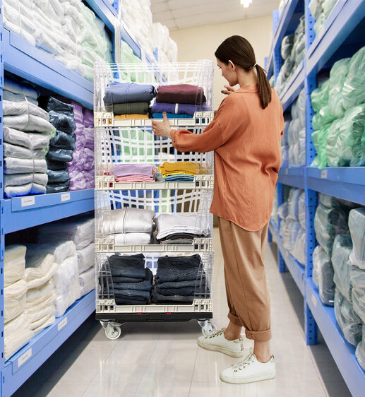 A woman selects products from storage shelving to restock Tosca Crystal Clear Crates displayed on a Tosca dolly, which offers easy movement for textile retail replenishment.