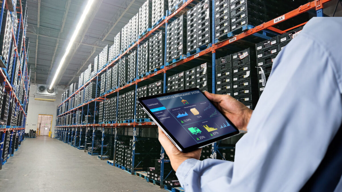A warehouse manager stands centered at the front of an aisle of tall racks storing reusable plastic containers (RPCs) stacked on top of pallets, while holding a tablet that displays a warehouse management system featuring a dashboard with graphics.