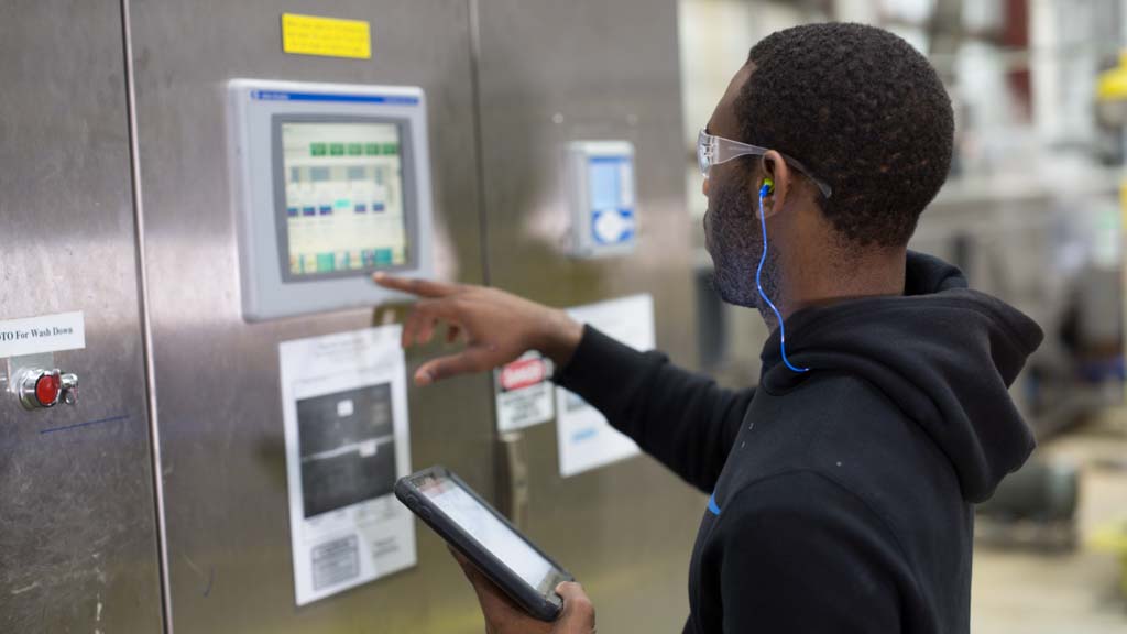 A man wearing ear plugs and safety eyeglasses touches a monitor displayed on a stainless-steel door, using technology to track the functionality of machinery in an industrial facility.