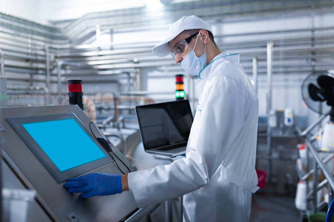 To ensure regulatory compliance, a man in a white coat, mask, and blue nitrile gloves touches a digital display with his left hand while holding a laptop in his right hand as he stands in a factory.