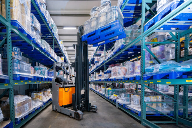 Warehouse scene with a worker operating a forklift to lift a stack of blue reusable plastic pallets loaded with packaged goods, demonstrating efficient storage and heavy-duty handling in a distribution facility
