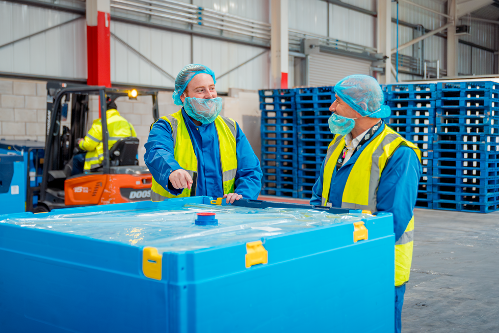Two workers in protective gear and high-visibility vests discussing near a large blue bulk reusable plastic container in a warehouse, with a forklift and stacked pallets in the background.