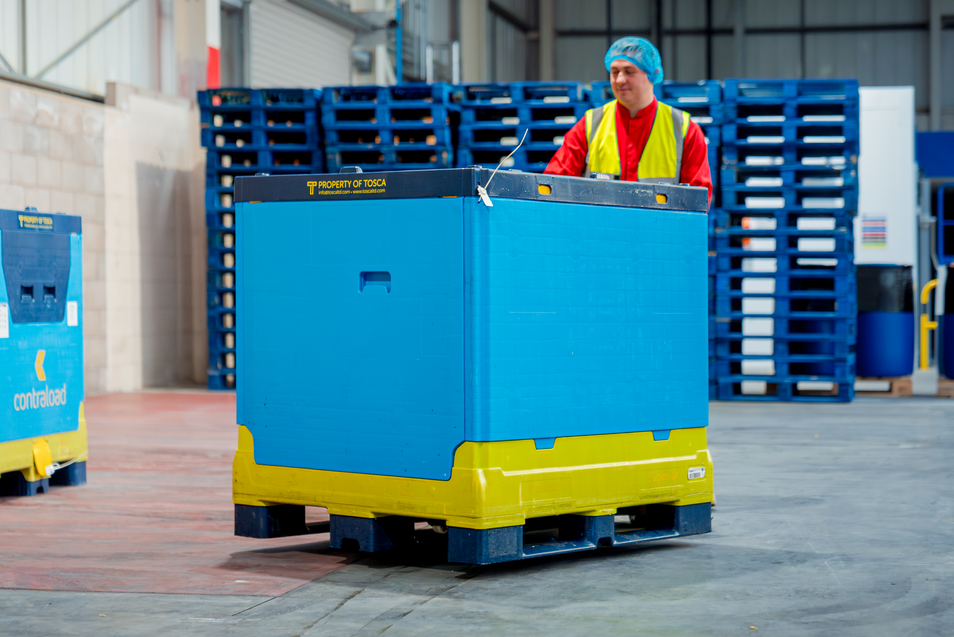 A worker in a red uniform and protective hairnet is handling a large blue and yellow reusable bulk container marked as “Property of Tosca.” The container is being moved within a warehouse setting, with stacks of blue pallets visible in the background.