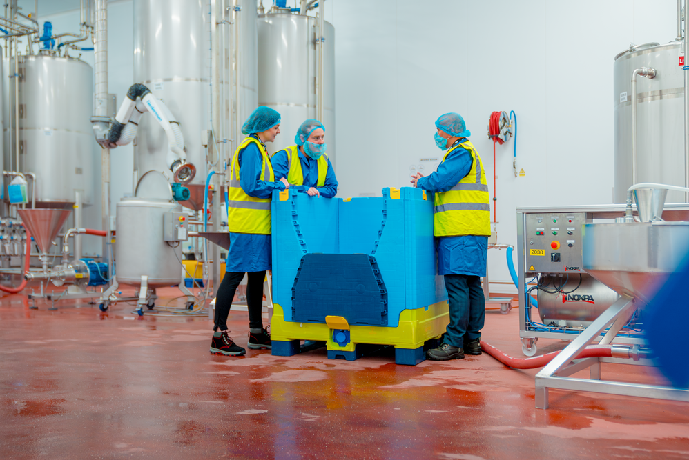 3 workers in high visibility clothing stand around a reusable plastic bulk container in discussion about something. The background is a clean warehouse facility