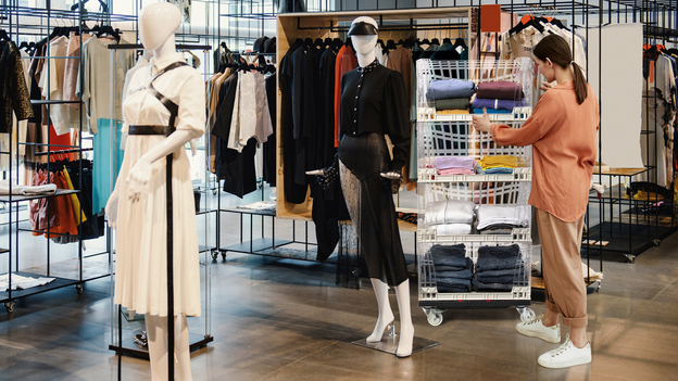 A woman shopping in a retail store browses clothing displayed in clear reusable plastic crates stacked on a portable Tosca plastic dolly.