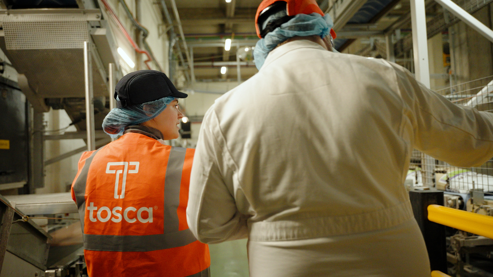 Two workers in a processing facility, one wearing a Tosca-branded high-visibility vest, engaged in discussion, highlighting collaboration and focus on safety and quality in an industrial environment.