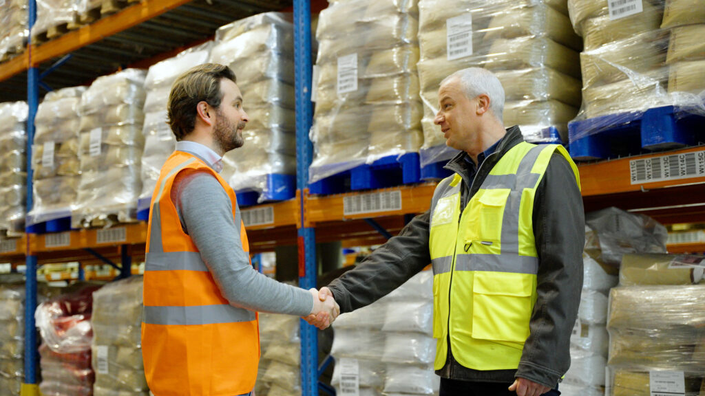 Two men in high-visibility vests are shaking hands in a warehouse setting, indicating a successful agreement or partnership. One man is wearing an orange vest, and the other is in a yellow vest. The background shows stacked pallets wrapped in protective film, emphasising the logistics and storage environment.