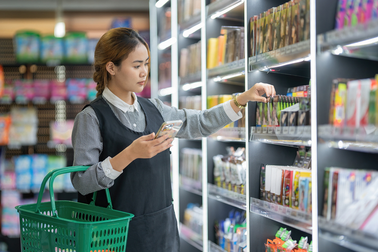 A woman selects products from a shelf while holding a reusable plastic shopping basket in a store aisle.