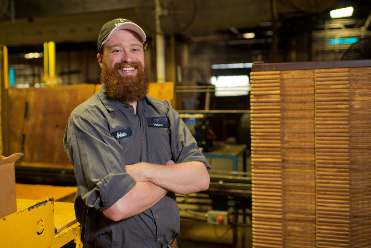 A Tosca cheese craftsman stands in front of stacks of wooden boards used to make Tosca wood cheese boxes inside a manufacturing facility.