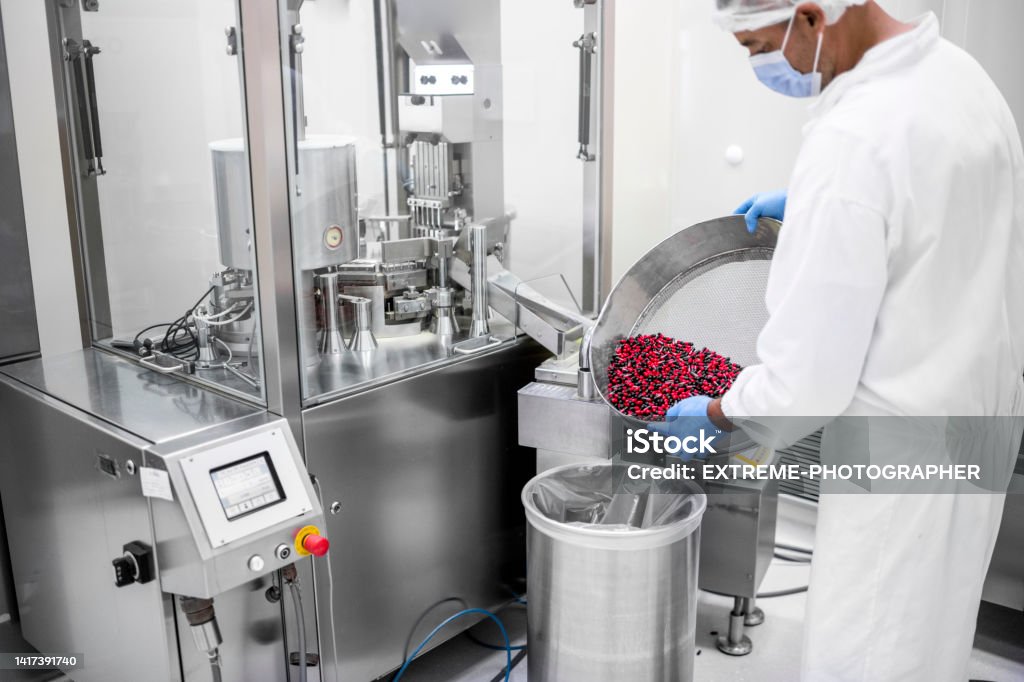 Pharmaceutical Technician Seen In A Laboratory While Holding A Big Bowl With Red And Black Pills During Working In A Laboratory In A Factory