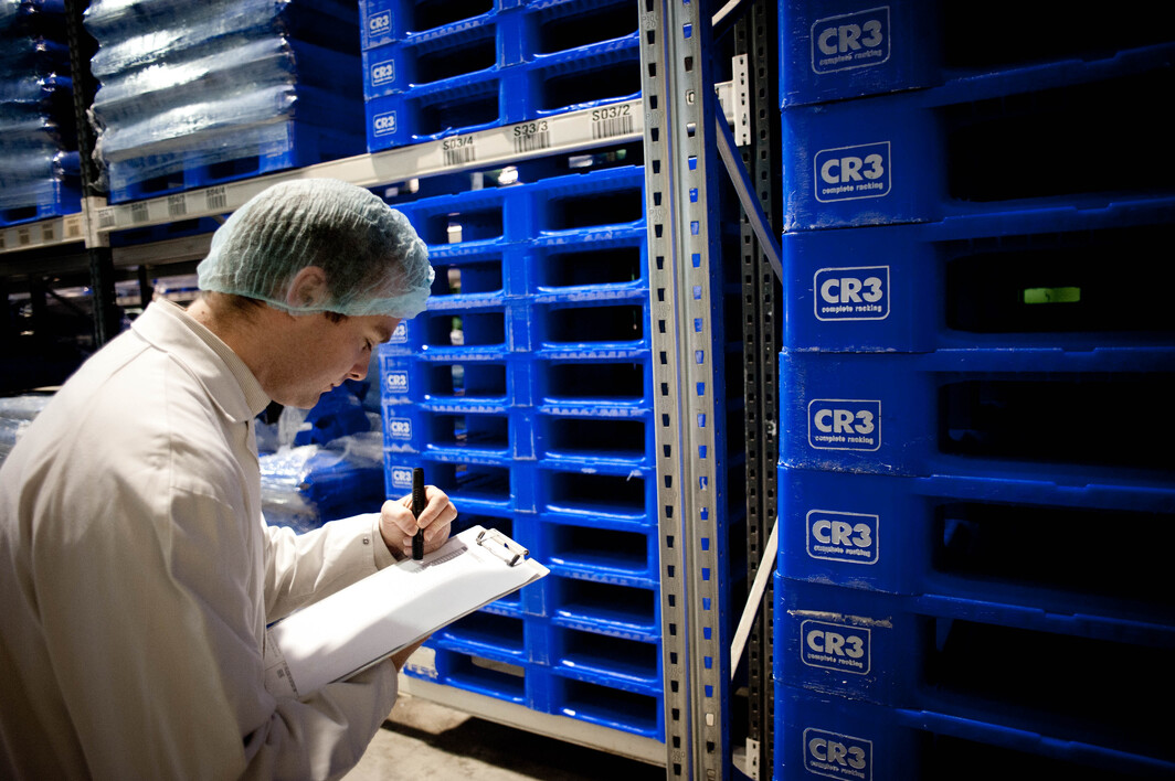 Worker in a cosmetics facility inspecting and taking notes near a stack of blue CR3 reusable plastic pallets, emphasising quality control and efficient storage in a hygienic environment.