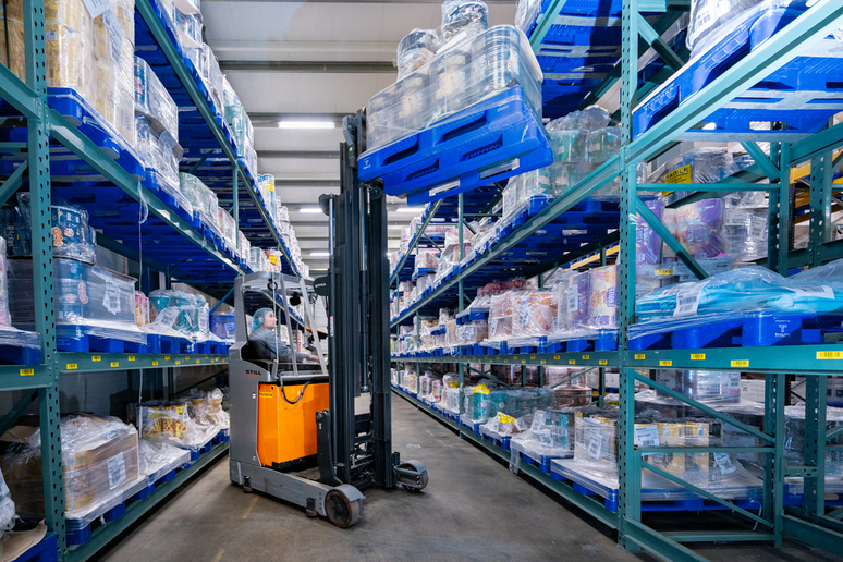 Warehouse scene with a worker operating a forklift to lift a stack of blue reusable plastic pallets loaded with packaged goods, demonstrating efficient storage and heavy-duty handling in a distribution facility