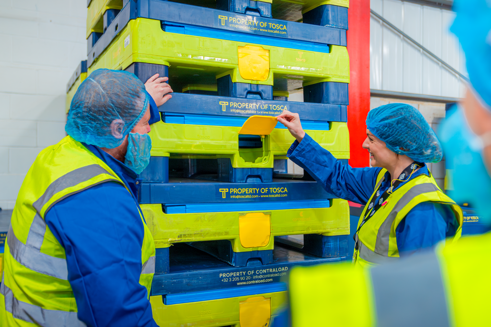 Two workers in protective clothing and high-visibility vests are inspecting a stack of blue and yellow reusable pallets branded with 