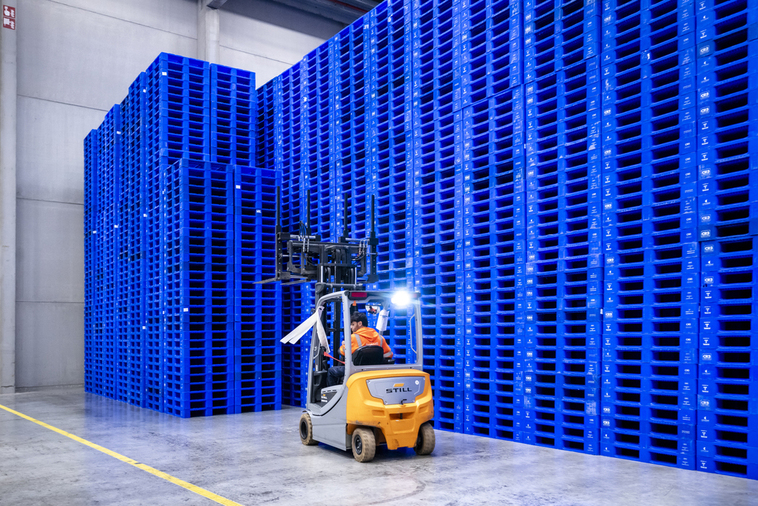 A forklift driver places blue plastic pallets onto stack of stable plastic pallets.
