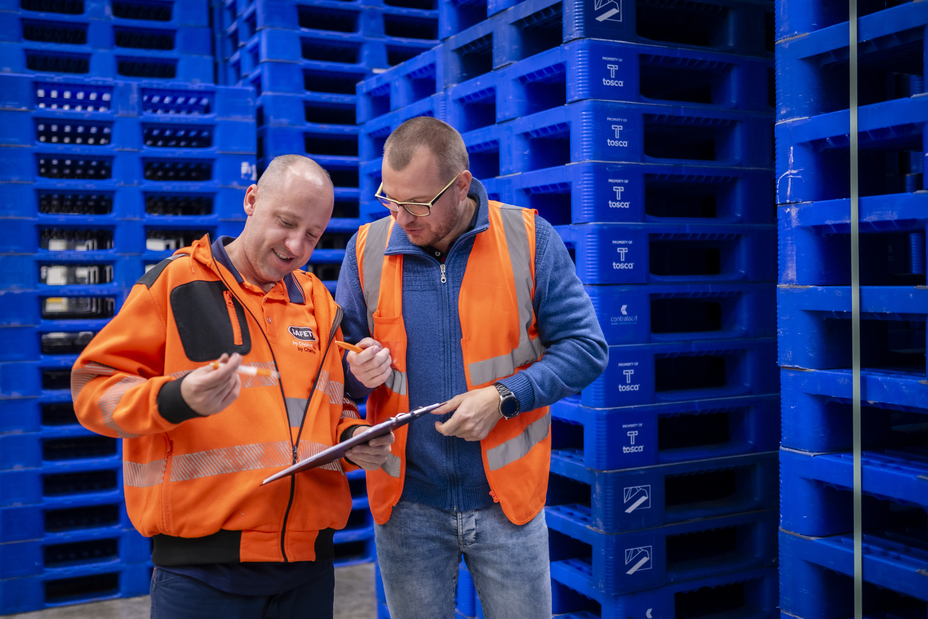 Two workers in orange high vis vests look at a clipboard and seem to be discussion. In the background is a stack of reusable plastic blue pallets.