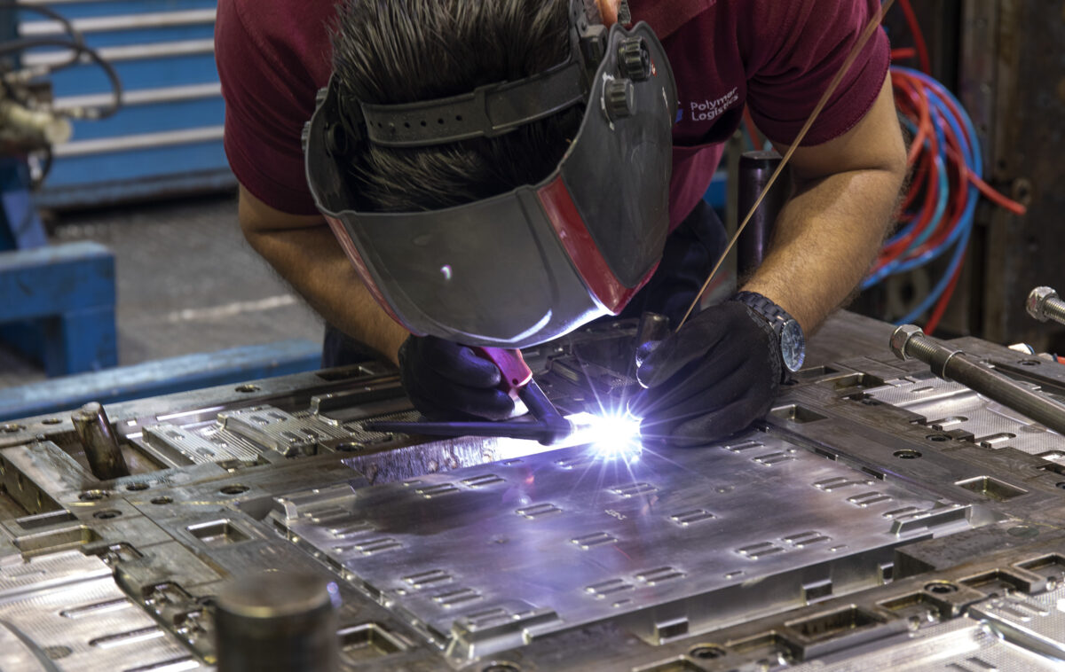 A worker wearing a protective welding mask and gloves is performing a precision welding task on a metal mold. Sparks and bright light are visible as the welding tool makes contact with the surface.
