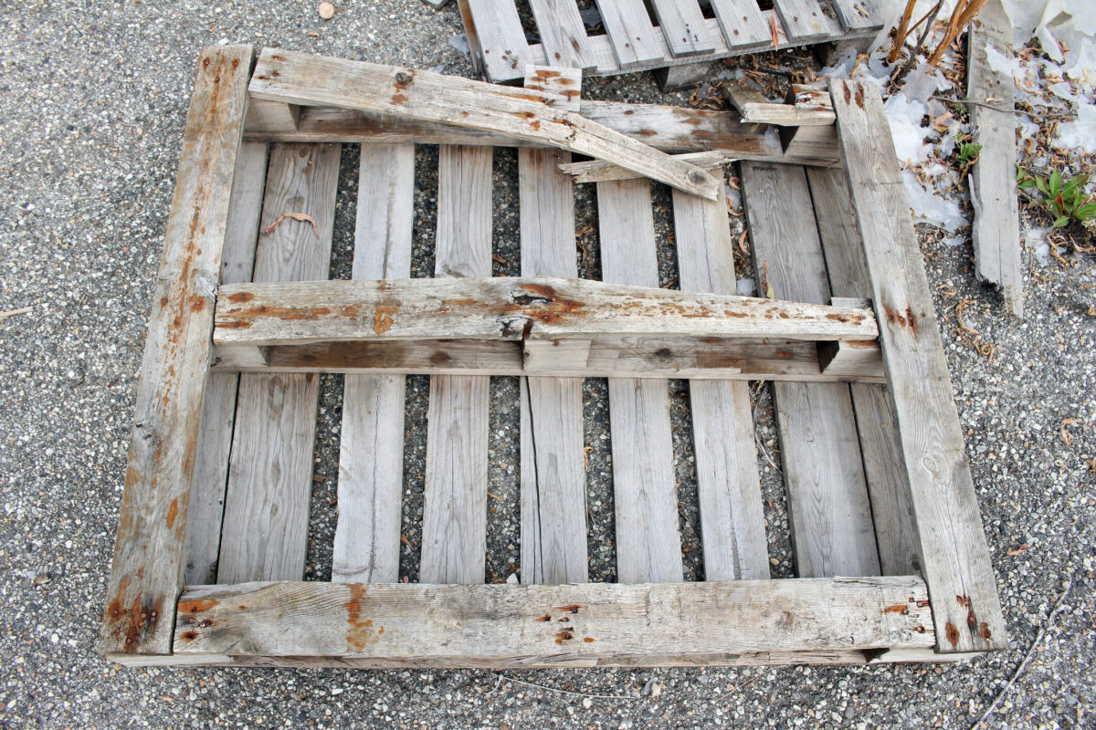 Weathered and damaged wood pallets with raised rusty nails, chipped slats, and broken boards. The lifespan of wooden pallets is much shorter compared to reusable plastic pallets.