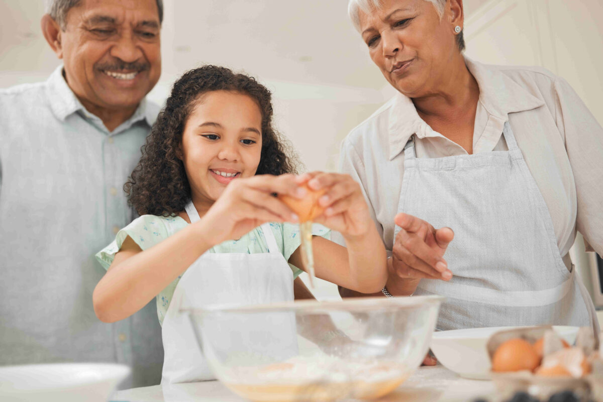 A little girl cracks open a brown egg over a bowl of flour as she prepares a meal with her grandparents in the kitchen.
