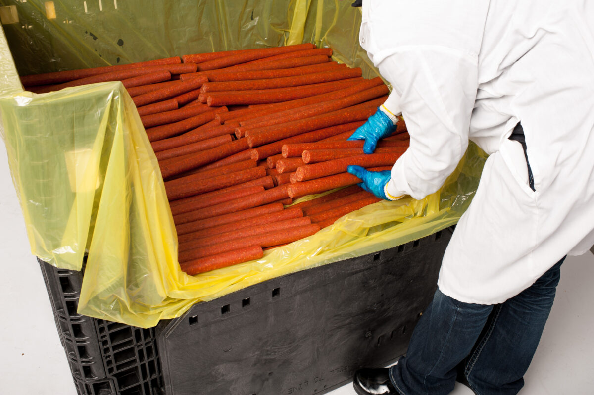 A worker is unloading meat sticks from a reusable Tosca General Purpose Bin, with the front folded down for easy access and less labor.