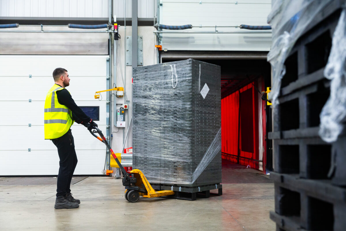A worker wearing a neon yellow safety vest uses hand trucks to move two tiers of folded crates, neatly stacked and secured to a plastic pallet in a Polymer Logistics environment.